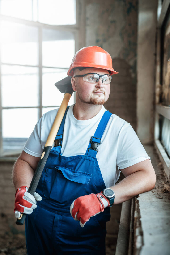 A tired worker stands near the window at a construction site. He is wearing a red safety helmet and he holds a sledgehammer for repairs in his hands.